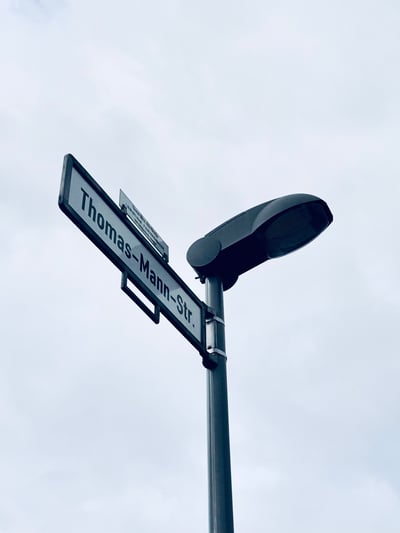 这是一张仰拍视角的街景照片，深灰色的路灯杆上固定着一块写有"Thomas-Mann-Str."的路牌，路牌旁还有一块小型说明标牌，背景是阴沉的浅灰色天空，画面风格简洁清冷，展现了德语区城市的一处普通街景。