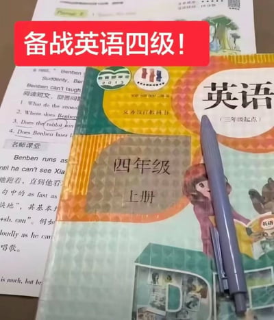 图片展示了一本小学英语教材和相关学习资料，上面覆盖着一个醒目的红色横幅，写着“备战英语四级！”。教材封面上清晰可见“英语（三年级起点）四年级上册”的字样，旁边还有一支笔放在教材上。这种用小学四年级课本准备大学英语四级考试的强烈反差，形成了一种幽默的调侃效果，暗示着准备不足或方法不当的学习状态。