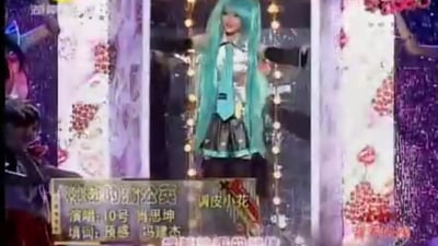 这是一张选秀类节目的视频截图，画面中间是10号选手肖思坤，他cos了虚拟歌手初音未来，身着标志性的蓝白配色初音服饰、佩戴蓝绿色长发假发，站在带有花卉装饰边框的舞台中进行演唱表演；画面下方的字幕显示，演唱曲目为《模糊的红公仔》，填词为预感、冯建杰，还有“调皮小花”的标识，画面左侧能看到其他在场人员，整体是将二次元cos形象与本土原创填词歌曲结合的舞台场景，带有跨次元的反差感。