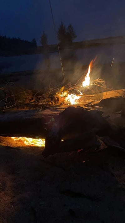 夜晚湖边的营火场景，火焰在黑暗中燃烧，照亮了周围的环境。篝火周围堆放着木柴，火焰上方升起袅袅青烟。远处可以看到平静的湖面和对岸的树木轮廓，天空呈现出深蓝色，显示出这是日落后的傍晚时分。整个场景营造出一种宁静而温暖的露营氛围。