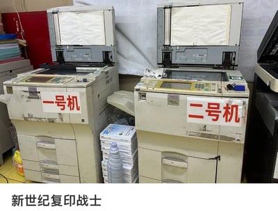 图片展示了两台并排摆放的老旧RICOH复印机，机身贴有红色醒目标签“一号机”和“二号机”，表面可见明显使用痕迹与污渍。机器顶部纸盒均处于打开状态，右侧机器操作面板上放置着一块白色抹布。设备旁堆放着复印纸、塑料瓶及清洁剂等耗材，背景为白色墙壁与红色柜子。底部配有文字“新世纪复印战士”，通过谐音梗化用经典动画《新世纪福音战士》名称，将办公场景中老旧但仍在服役的复印机幽默比作“战士”。