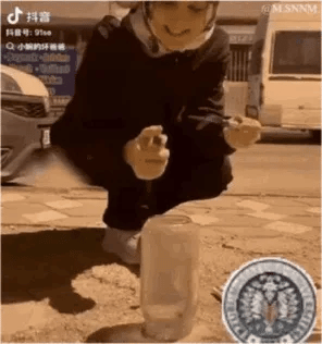 这是一段动态GIF，展示了户外的搞笑失误场景：一名男子蹲在地面的塑料杯前，手持类似点火器的道具，似乎要点燃杯中的物品，结果在操作过程中脚下突然踩空，整个人掉进了地面的一个坑洞，最后塑料杯中冒出烟雾，男子消失在画面里，整个过程充满意外感，让人忍俊不禁。