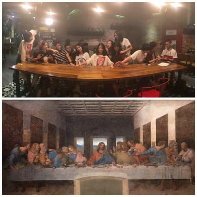这是一张上下分栏的对比图，上图是一群年轻人在室内聚会场所，精准复刻达芬奇经典名画《最后的晚餐》的人物构图与姿态，围坐在长桌旁摆出对应姿势，氛围轻松欢乐；下图是列奥纳多·达·芬奇的原作《最后的晚餐》，呈现耶稣与十二门徒共进晚餐的经典宗教场景。两者构图高度对应，是一次充满趣味的名画创意模仿创作。