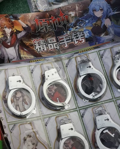 图片展示了一款原神主题的周边商品，包装上印有游戏内知名角色（红发的迪卢克与蓝发的优菈），主打产品为银色手铐造型的饰品，手铐圆环内侧印有游戏角色图像。商品名称“原神 精品手铐”直接印在包装上方，整体设计将热门二次元游戏IP与非常规的手铐造型结合，形成强烈的视觉反差，引发网友“buff叠满”的调侃，因其组合方式新奇且略带幽默。