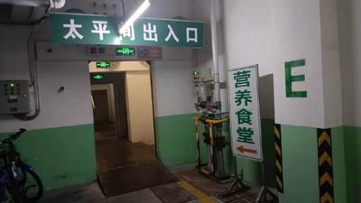 这是一张在医院内部拍摄的实景照片，画面上方悬挂着绿色的「太平间出入口」标识牌，下方门口亮着绿色的安全出口指示灯，门内是光线昏暗的通道；画面右侧立着一个指向左方的「营养食堂」指示牌，旁边的墙面上标有字母「E」，左下角还停放着一辆自行车。场景的荒诞感来源于太平间与食堂这两个功能完全不同的区域，指示牌位置相近，容易引发观者的趣味联想。