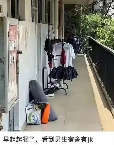 这张图片展示了男生宿舍的走廊场景，走廊的衣架上挂着几件衣物，由于白色上衣和黑色百褶裙的摆放位置，看起来像是一套JK制服，旁边地面还放着鞋子、背包和购物袋等杂物。图片下方配文“早起起猛了，看到男生宿舍有jk”，利用视觉错觉制造出反差感，形成搞笑的乌龙效果，实际只是普通衣物的组合摆放造成的误会，并没有真的JK制服出现在男生宿舍。