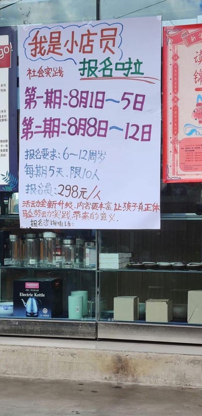 这是一张张贴在店铺玻璃上的手写招募海报，面向6-12周岁儿童推出「我是小店员」社会实践活动，活动分为两期：第一期8月1日-5日，第二期8月8日-12日，每期开展5天，每期限10人，报名费298元/人。海报说明活动已全新升级，内容更丰富，能让孩子真正体验劳动实践的意义，下方预留了报名咨询电话的填写位置。海报下方的玻璃展示柜中，陈列着水杯、电热水壶等商品。