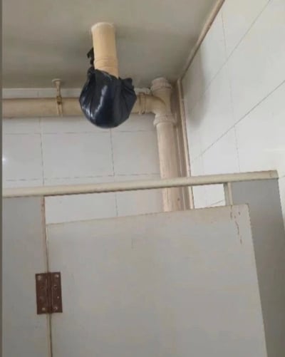 这是一张公共卫生间的实拍图，画面里天花板下方的下水管道出现破损，人们没有对管道进行正规维修，仅用黑色塑料袋套在管道破损处承接漏出的污水，下方可见卫生间的隔断挡板，这种敷衍的临时处理方式搭配对应的配文十分具有讽刺幽默感，吐槽人们仿佛只要把脏东西用塑料袋装起来就可以忽略其存在的心理。