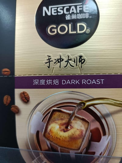 雀巢咖啡金牌系列的手冲大师深度烘焙产品包装特写。包装主体为金色拉丝质感，顶部有黑色圆形标志，内有白色'NESCAFE 雀巢咖啡'和金色'GOLD'字样。中间偏下位置有中文'手冲大师'手写风格文字，下方紫色条带标注'深度烘焙 DARK ROAST'。底部图案展示一杯咖啡正被倒入，旁边散落几颗咖啡豆，整体设计凸显高端和烘焙深度。