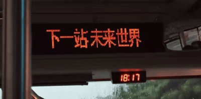 这是一张在公交车内拍摄的静态照片，画面主体是车内上方的电子站点显示屏，以橙色点阵字体清晰显示“下一站未来世界”，充满了科幻感与浪漫的想象色彩；显示屏下方的电子时钟显示时间为18:17，此时天色看起来偏暗，结合前挡风玻璃上的雨水痕迹，能看出当时是雨天；窗外是模糊的绿色植被，整体场景营造出一种仿佛乘坐公交就能前往充满憧憬的未来世界的治愈氛围，“未来世界”并非真实的公交站点，是带有创意加工的内容。