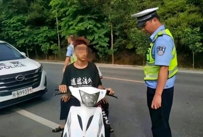 这是一张真实的现实场景抓拍照片，画面中一名面部被马赛克处理的男子坐在白色电动车上，他身着印有「逍遥法外」字样的黑色T恤，正被一名身着反光背心与警服的交警拦下，交警低头看向男子，似乎正在进行执法检查。在男子身后还有另一名交警，画面左侧停着一辆带有「警察」「POLICE」标识的警用轿车，车牌为「桂N·N9939警」，背景是道路旁的茂密绿植。男子T恤上的文字与被交警拦截的现实场景形成强烈的反差，制造出极具戏剧性的搞笑效果，是偶然发生的趣味巧合场景。