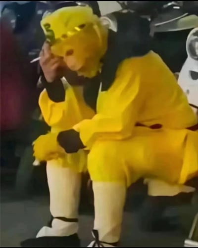 图片中显示一个穿着黄色猴子全套服装的人，正坐在椅子上，左手拿着一根香烟靠近嘴边，右手支撑着头，呈现出一种沉思或疲惫的姿态。人物穿着黄色连体猴子服，戴着黄色头套，头套上有简单的面部特征。背景模糊，可以看到一些模糊的物体和可能的其他人物，但主体清晰可见。这张图片没有明显的双关、谐音梗或暗示内容。