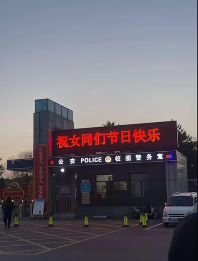 图片展示了一座校园警务室的外观，拍摄于黄昏或黎明时分，天空呈现淡蓝色。警务室建筑为深色，顶部有一块大型红色LED显示屏，醒目地滚动显示“祝女同胞们节日快乐”的字样。建筑正面悬挂着“公安 POLICE 校园警务室”的标牌，标牌右侧有红蓝警灯。警务室门口有玻璃门和窗户，窗内隐约可见室内陈设。门前的空地上摆放着多个黄色交通锥，一辆白色面包车停放在右侧，左侧有一人正步行经过。建筑左侧有红色对联，上面写有“三全育人 有生态人 更卓越”等字样，整体场景温馨，体现了节日祝福的氛围。