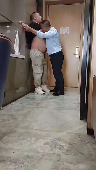 在一个类似酒店卫生间的狭小空间里，一位穿着浅蓝色衬衫黑裤子的男子，将一位上衣掀起露出大肚子、穿着卡其色裤子的胖男子壁咚在墙边，胖男子被抵在墙面，头部后仰，手臂抬起，表情看起来有些无奈或惊讶，画面充满搞笑感，明显是朋友间的玩闹整活场景。