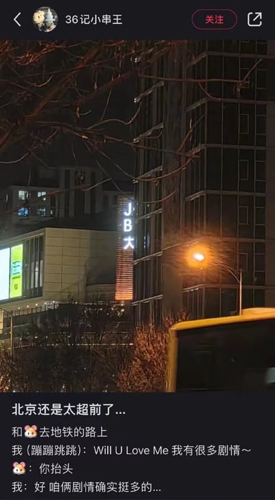 这是一张社交媒体截图，展示了北京夜晚的街景照片。照片中可以看到一栋建筑物，其外墙上有一个亮着灯的招牌，显示“JB大”字样。照片下方配有一段文字，内容是用户与朋友去地铁路上的对话：用户蹦蹦跳跳地说“Will U Love Me 我有很多剧情～”，朋友让其抬头，用户看到招牌后回应“好 咱俩剧情确实挺多的...”，整体氛围轻松幽默，带有调侃意味。