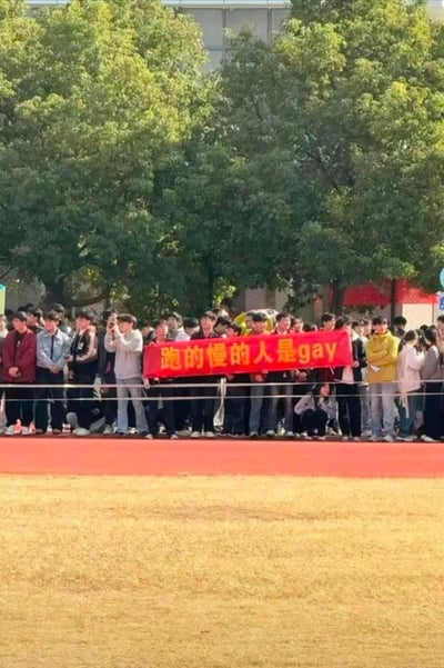 校园活动现场，一群学生举着写有'跑的慢的人是gay'的红色横幅，周围有许多围观学生。该标语因使用性取向相关词汇作为调侃内容引发争议，反映了校园文化中有时出现的不当玩笑现象。