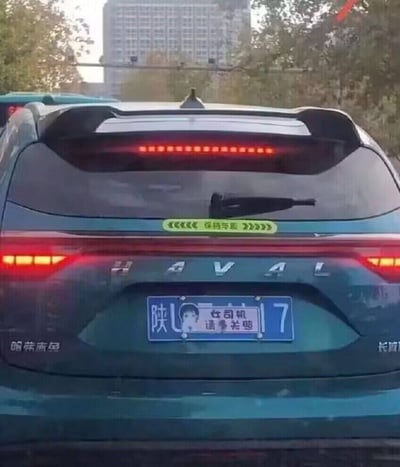 照片拍摄于城市道路上，视角来自后车，主体是一辆蓝绿色哈弗SUV的尾部。车辆顶部的高位刹车灯和两侧尾灯呈红色点亮状态，后挡风玻璃上装有雨刷。玻璃下方贴有一条黄绿色长条提示贴，两侧分别是向左和向右的箭头，中间写着“保持车距”。车尾号牌为蓝色，左侧可见“陕U”，右侧可见数字“7”，号牌中间区域被一张白色贴纸覆盖；贴纸上有一个黑色短发的卡通女孩形象，并配有“女司机 请多关照”两行字。该贴纸用自嘲和幽默的方式提醒后车驾驶员保持耐心与车距。背景中可以看到树木、远处的高层建筑物以及另一辆车的局部，整体像是在傍晚或白天光线柔和时的道路随拍。画面没有明显事故或冲突，主要趣味点来自车尾标语与驾驶身份标签。