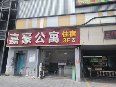 这是一张城市临街底商的实拍图，画面主体为一楼嘉豪公寓的入口区域：入口上方悬挂红底招牌，左侧是白色立体大字“嘉豪公寓”，右侧配有黄色字体“住宿 3F”及向上双箭头，提示住宿服务位于建筑三层。入口为透明玻璃门，门上贴有“欢迎光临”标识与联系电话，门口停放着一辆电动自行车，左侧放置有黑色铁马护栏，右侧墙面贴有带卡通头像的二维码，墙边立有绿色共享充电宝装置。入口右侧是停车场通行通道，可看到通道内部的“壹味饺”商铺标识与停放的车辆，建筑外墙上部为黄白相间的条纹装饰，上方还露出部分水果主题的广告牌，整体是生活化的城市街景场景。