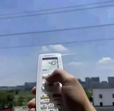 这是一张利用视觉错位创作的趣味实拍图，画面中一只手握着温度设定为40℃的空调遥控器，朝向窗外的蓝天与城市远景，制造出仿佛可以用空调遥控器直接调节地球户外气温的搞笑效果，搭配相关配文形成玩梗内容，契合夏季高温天气的大众热议语境。