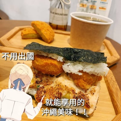 图片展示了一盘创意日式料理，主体是一个由米饭、海苔、炸物堆叠而成的食物，放置在木质托盘上。托盘旁还有两块炸薯饼和一杯纸质杯装饮品，背景有一个玻璃瓶装饮品和包装食品。图片左侧有一个动漫风格的人物贴纸，人物侧身指向食物，脸上带着微笑。图片上覆盖有文字“不用出國 就能享用的 沖繩美味！！”，突出宣传无需出国即可品尝到冲绳风味的美食。整体画面色彩鲜亮，食物看起来金黄诱人，营造出轻松愉悦的美食分享氛围。