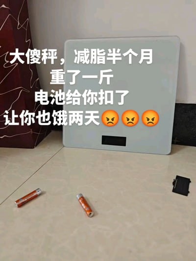 这是一张实拍的生活搞笑梗图，画面中电子体重秤靠在墙边，其电池已经被拆下，两节电池和电池盖散落在前方的浅色瓷砖地面上。图上叠加了白色配文，内容为“大傻秤，减脂半个月重了一斤，电池给你扣了，让你也饿两天”，还搭配了三个愤怒的表情，十分生动地展现了减肥遇到瓶颈、体重不降反升的当事人迁怒体重秤的搞笑又无奈的心态。