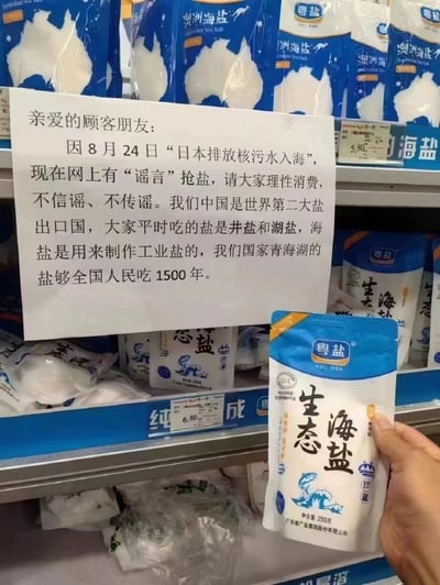 这是一张在国内超市食盐货架区域拍摄的实拍图，货架上陈列着粤盐品牌的澳洲海盐、生态海盐等多款食盐产品，货架前方显著位置贴着一张手写告示。告示针对2023年8月24日日本排放核污水入海后网络上出现的抢盐谣言，向顾客发出呼吁，告知大家要理性消费，不信谣不传谣，同时科普了中国的食盐供应情况：中国是世界第二大盐出口国，民众日常食用的盐多为井盐和湖盐，海盐多用于工业生产，青海湖的盐储备量足够全国人民食用1500年，以此打消民众的抢盐顾虑。画面右下角有一只手拿着一袋粤盐生态海盐，清晰展示了产品的包装细节。