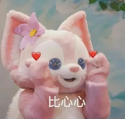 图片中是迪士尼粉色小狐狸角色玲娜贝儿的毛绒玩偶特写，它有着粉白渐变的毛发，左耳佩戴一朵粉色花朵发饰（中心有黄色花蕊），一双水汪汪的蓝色大眼睛格外灵动。玩偶正用两只毛茸茸的前爪做出比心手势，眼睛两侧各有一个红色爱心图案特效，下方配有白色文字“比心心”，整体画面软萌治愈，充满可爱气息。