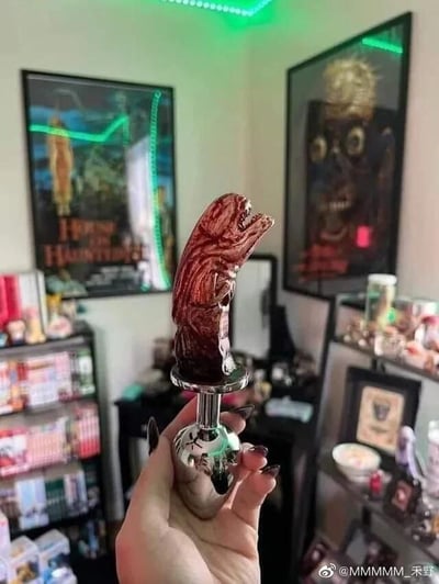 一只涂着黑色美甲的手握着一个造型诡异的红色收藏品，该收藏品呈现出类似怪物张开嘴巴的恐怖形态。背景是一个充满暗黑风格的房间，墙上挂着《猛鬼街》等经典恐怖电影海报，绿色LED灯带勾勒出墙面轮廓，营造出阴森氛围。房间内摆放着多层展示架，上面陈列着各种动漫手办和收藏品，整体呈现出浓厚的恐怖文化爱好者的私人空间特色。