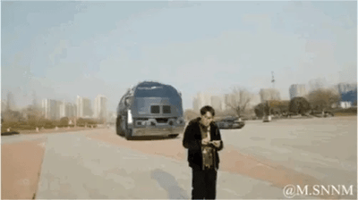 这是一段创意合成的动态GIF，开篇呈现一名男子站在城市户外道路旁看手机的场景，随后画面中凭空出现《红色警戒》风格的大型载具，载具随即变形展开，逐步生成红警系列游戏中的核心基地建筑、采矿场、战车工厂等设施，同时还有配套的坦克单位同步出现，将现实城市道路场景与经典RTS游戏《红色警戒》的建造系统进行创意融合，以视觉合成的方式模拟出在现实中搭建红警基地的趣味过程，充满怀旧感与创意性，勾起玩家对经典游戏的回忆。