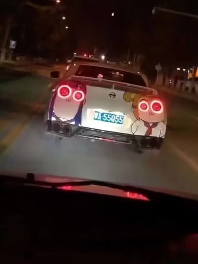 这是夜间城市道路场景下，从后车驾驶位向前拍摄的前车车尾照片。画面中的前车为白色日产轿车，车尾车牌为陕A·55Q55，车牌左右两侧分别张贴了动漫《POP子和PIPI美的日常》中的两个主角POP子、PIPI美的半身卡通贴纸，贴纸的眼睛位置恰好和车辆的红色刹车灯位置重合，当刹车灯亮起时，刚好形成两个卡通角色睁着明亮红色发光大眼睛的趣味视觉效果，画面底部可见后车的挡风玻璃边缘及部件。