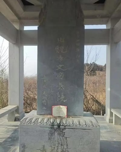 这是一张在碑亭内拍摄的北魏孝文帝长陵纪念碑照片。碑身为灰色石材，正面镌刻着金色的“北魏孝文帝长陵”字样，右上方标注有“二〇一四年八月”的题字，左侧则是“孟津县人民政府立”的落款。碑座带有精致的莲花瓣样式雕刻，碑的正前方放置了一个带有红边的说明牌。碑亭由浅色石柱和横梁构成，亭外可见枯黄的草木与远处的低矮山丘，整体氛围古朴肃穆，完整呈现了这座帝王陵寝纪念设施的样貌与周边环境。