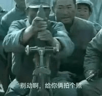 图片展示了几名穿着军装的士兵，其中一人正双手持枪并将枪托抵在 shoulder 上，做出类似拍照的姿势对准前方，旁边有其他士兵围观。画面底部配有文字“别动啊，给你俩拍个照”，通过将步枪模仿成相机的创意，形成了幽默的反差效果，原本严肃的军事场景变得诙谐有趣。