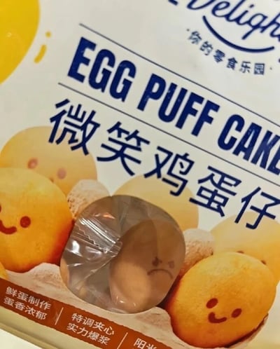 这是一张零食包装袋的近距离实拍图。产品名为“微笑鸡蛋仔”，包装上画着多枚带有红色微笑表情的鸡蛋仔，并配有“鲜蛋制作 蛋香浓郁”“特调夹心 实力爆浆”等宣传语。包装中央有一个透明窗口，可以看到里面真实的圆形鸡蛋仔。由于糕点本身的色泽、阴影以及塑料包装的褶皱相互叠加，实物表面恰好形成两个类似眼睛的深色小点和一条下弯的弧线，看起来像在皱眉、撇嘴或难过地哭丧着脸。它与包装上整齐可爱的微笑图案形成强烈反差，制造出“名义上微笑，实物却高兴不起来”的幽默效果。发布者配文仅一个“🙁”表情，进一步强化了这枚鸡蛋仔的沮丧感。