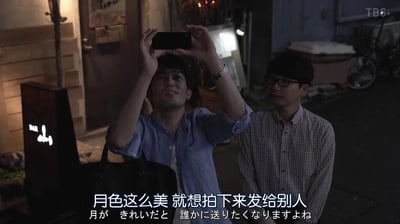 这是一张日本影视的横版截图，场景设定在夜晚的户外小巷中，画面左侧有一个带有“BAR”字样的发光灯牌，背景里能看到关闭的门和停放的自行车，营造出静谧的夜晚氛围。画面中有两位男性，左侧的男子身着蓝色衬衫，双手举着手机，脸上带着柔和的笑意，眼神中透着憧憬，似乎正在拍摄夜空中的月亮；右侧的男子戴着眼镜，穿着格子衬衫，站在一旁，神情温和，正看向举手机的男子，仿佛在倾听对方的话语。画面下方配有中日双语字幕，中文内容为“月色这么美 就想拍下来发给别人”，对应的日文是“月が きれいだと 誰かに送りたくなりますよね”，传递出看到美好事物时，想要分享给在意之人的细腻、温柔的情绪，是一种含蓄表达思念与在意的方式。