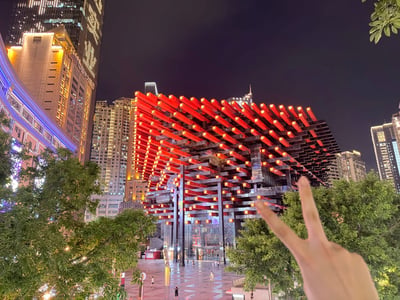 这是一张夜间拍摄的城市实景照片，画面主体是重庆地标建筑国泰艺术中心，该建筑造型极具辨识度，外观由大量向外穿插的红色构件堆叠而成，构件端部点缀有亮黄色灯光，视觉效果十分醒目。建筑周边是重庆渝中区的高层商务楼与住宅楼，均已亮起夜间照明，展现出繁华的都市夜景。前景右下角有一只真人的手比出V字剪刀手手势，似乎是拍摄者在打卡留念，画面两侧有绿色的行道树，下方是开阔的城市广场，广场上有少量行人正在活动。