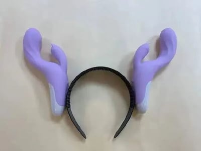 这是一张静态单图，属于原始拍摄图，画面主体是一个黑色的普通发箍，发箍的两侧安装了紫色的装饰，该装饰的造型高度模仿某类女性情趣用品，仅在底部做了浅灰色的处理，试图伪装成类似鹿角的形态，是一款充满恶搞趣味的整蛊道具，通过造型的相似性制造出荒诞搞笑的视觉效果，图中没有文字内容，也不存在双关、谐音梗等元素。