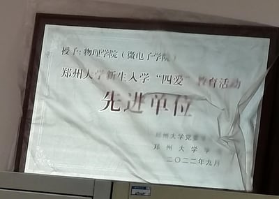 图片展示了郑州大学颁发的一份荣誉证书，内容为授予物理学院（微电子学院）‘郑州大学新生入学‘四爱’教育活动先进单位’称号，落款为郑州大学党委和郑州大学，日期为二〇二二年九月。证书被装裱在相框中，表面覆盖的塑料薄膜有轻微褶皱。