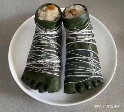 这是一张展示创意美食的实拍图，盘中摆放着两个被塑造成人类脚丫模样的粽子。粽叶被折叠出脚趾的形状包裹住粽身，粽身用白色线绳交叉缠绕固定，顶部被切开，露出内部的糯米和馅料，将传统端午美食粽子做出了搞怪有趣的新奇造型，视觉效果诙谐，充满创意。