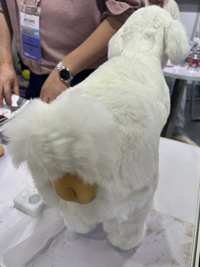 图片展示了在 AI EXPO 展会现场的一个白色毛绒狗玩具，玩具的尾部有一个突出的圆形黄色部件。背景中可以看到一位穿着粉色衬衫的工作人员，佩戴着带有 'AI EXPO' 字样的蓝色参展证件和一块黑色手表。桌子上还放着一个小型白色电子设备，周围有瓶装水等展会常见物品。整体场景呈现了科技展会中展示可爱毛绒玩具的有趣对比画面。