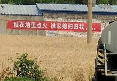 这是一张户外实拍的乡村场景图，画面主体是一片成熟待收的金黄色麦田，麦田左前方有一小丛绿色植物，右侧露出一辆罐车的部分车身结构；背景是带有蓝灰色彩钢屋顶的砖制房屋，房屋外墙上悬挂着一条醒目的红色宣传横幅，横幅上印着“谁在地里点火 谁家媳妇归我”的戏谑式标语，用夸张、搞笑的表述来警示村民不要在田间焚烧秸秆，以此避免引发火灾、污染环境，这种接地气的宣传方式旨在加深村民的记忆，达到更好的禁烧宣传效果。
