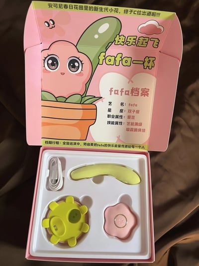 图片展示了一款来自「安可尼春日花园」的粉色产品包装盒，盒身印有卡通小花角色「fafa」的可爱形象——圆滚滚的粉色花身、绿色叶片，搭配超大萌眼和微笑表情。包装盒顶部文字写着「安可尼春日花园里的新生代小花，终于C位出道啦!!!」，侧面标注「快乐起飞 fafa一杯」。盒内「fafa档案」详细介绍其艺名fafa、双子座、爱豆职业属性及「艺能满级 吸震俱佳」的技能，还提到正在全国巡演传递快乐能量。打开的盒内可见白色数据线、黄色弧形配件、黄色花形部件及带「K」标识的粉色花形部件，整体设计以粉绿为主色调，充满少女心与元气感，主打可爱偶像IP周边概念。