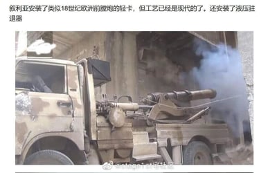 这张图片展示了一辆经过本土化改装的军用卡车，卡车采用荒漠迷彩涂装，后部搭载了一门外观酷似18世纪欧洲前膛炮的火炮，此时火炮正处于发射状态，炮口冒出明显的烟雾。根据图中文字说明，这门火炮虽然外形复古，但实际采用了现代工艺制造，还配备了液压驻退器来缓冲火炮发射时的后坐力，让这种复古外观的火炮能适配现代卡车平台，车辆停在一处简陋的混凝土建筑旁，整体场景显示这是在实际的作业或作战环境中。