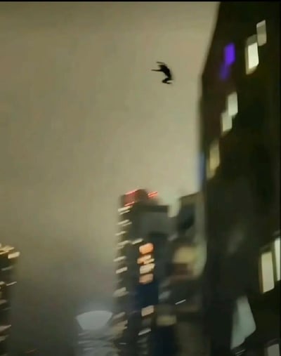 这是一张竖屏的视频截图，画面呈现夜晚的都市高楼群，背景中高楼的灯光点亮，整体画面有些模糊。在画面上方的空中，有一个黑色的人形剪影，正处于半空之中，看起来像是从高楼坠落或是进行高空跳跃的动作，营造出紧张惊险的氛围。