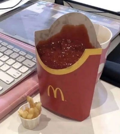 图片中展示了一个麦当劳红色薯条盒，盒内装满了深红色的番茄酱，几乎快要溢出。薯条盒旁边放着一个白色的小纸杯，里面只装着寥寥几根薯条。背景中可见粉色的键盘、平板电脑以及带有粉色圆点图案的白色杯子，整体场景像是在办公桌或学习桌上。画面的幽默之处在于颠覆了常规的薯条与番茄酱搭配比例——本该装薯条的盒子被番茄酱占据，而薯条却少得可怜，形成荒诞又搞笑的视觉对比。