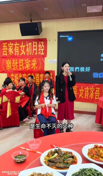 图片展示了一场特殊的庆祝活动现场，背景悬挂多条红色横幅，上面写有“吾家有女初月经”“庆祝朱府喜来”等醒目文字。一位身着黑色外套红色连衣裙的女性手持麦克风站立发言，前方坐着一位系红色围巾的少女，周围有多位中老年亲友围观就座。前景餐桌上摆放着多道菜肴，包括炒菜、炖菜等。屏幕上显示“我们很好 花多”的文字，画面底部叠加字幕“是生命不灭的火种”。整个场景因将月经初潮作为公开庆祝事件而显得较为罕见和具有争议性。
图片底部水印注明“剧情演绎 仅供娱乐 无不良引导 请勿过度解读”，表明内容为虚构演绎，非真实生活场景。