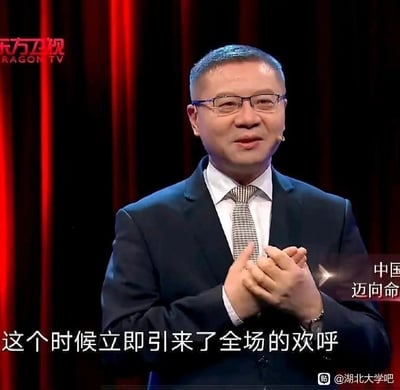 一位穿着深蓝色西装、白色衬衫和格子领带的中年男性，站在红色幕布背景前，双手合十鼓掌。画面左上角有东方卫视的红色台标，底部有文字“这个时候立即引来了全场的欢呼”，右下角有水印“@湖北大学吧”。