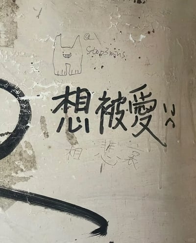 这是一张拍摄城市剥落墙面的照片，墙面上有创作者留下的手绘涂鸦：上方是一只咧嘴露齿的简笔小猫，旁侧标注英文“Stop3mins.”；中间是醒目的手写繁体文字“想被愛”，字体设计将“想”拆分为“相”与“心”，还在文字两侧添加了小表情符号；下方有浅色的手写小字“想悲哀”。墙面本身的剥落、斑驳质感，与直白又带着落寞的文字内容相呼应，传递出渴望被爱却又夹杂着悲哀的细腻情绪。
