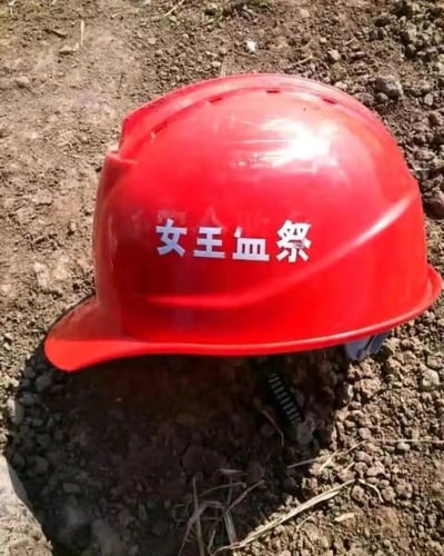 这是一张原始拍摄的静态图，画面主体是一顶放置在混杂小石子的泥土地面上的红色工地安全帽，安全帽正面印有醒目的白色文字“女王血祭”，和工地安全帽的实用属性形成强烈反差，带有玩梗的趣味感，安全帽下方能看到部分固定用的卡扣结构，整体场景看起来是户外工地或类似的户外场地。