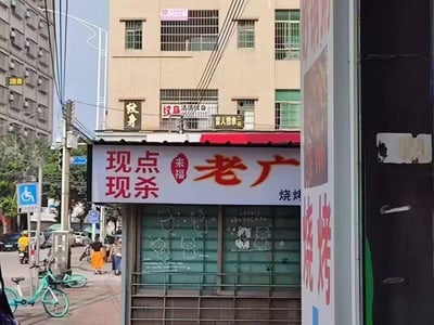这是一张城市街头实拍图，画面主体是一家名为“老广”的烧烤店，招牌醒目地写着“现点现杀 来福 老广 烧烤”，店铺的卷帘门上绘制着可爱的卡通猫咪图案，标注有“Lovely BabyCat”字样。背景的建筑外墙上挂着纹身店、盲人推拿店的招牌，街道上有行人路过，还停放着共享单车，旁边设有无障碍设施标识，整体呈现出充满生活气息的城市街角烟火场景，没有双关或谐音梗内容。