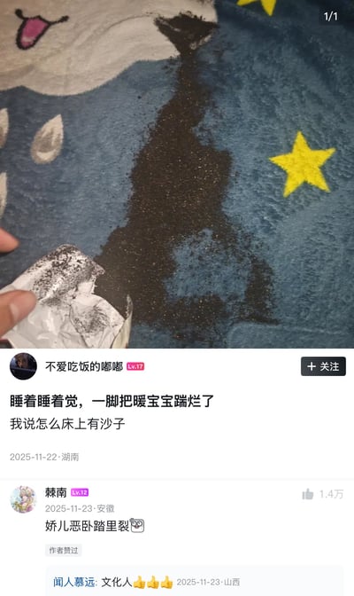 图片上半部分为实拍场景：蓝色带有黄色星星图案的毯子上，一个暖宝宝包装袋被撕裂，黑色颗粒状内容物大量泄漏并形成长条状污渍，一只手正捏着破损的包装袋边缘。下半部分为社交媒体帖子界面，用户“不爱吃饭的嘟嘟”配文描述“睡着睡着觉，一脚把暖宝宝踹烂了 我说怎么床上有沙子”，并显示发布于2025年11月22日湖南。下方有用户“棘南”的评论“娇儿恶卧踏里裂”（改编自杜甫《茅屋为秋风所破歌》中“娇儿恶卧踏里裂”），获1.4万点赞。